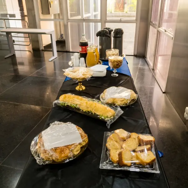 Mesa longa coberta com toalha preta, organizada como buffet com diversos tipos de pães, bolos e salgados cobertos por filme plástico, além de garrafas térmicas, suco e refrigerante ao fundo.